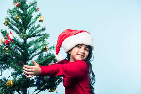 cheerful Indian girl child with santa hat posing or holding golden decorative balls over white or red or blue background on christmasの写真素材