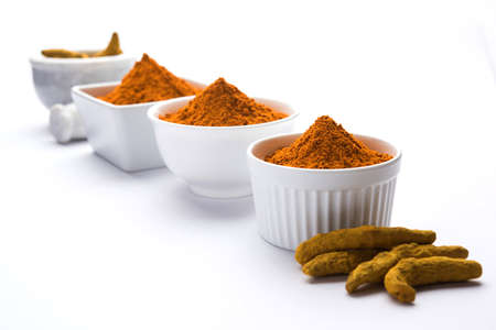 Turmeric powder in ceramic bowl with raw dried turmeric over plain background