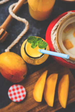 Mango Lassi or smoothie in big glass or small bottles with curd, cut fruit pieces and blender. Moody background, selective focusの写真素材