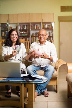 Senior Indian/asian couple accounting, doing home finance and checking bills with laptop, calculator and money while sitting on sofa/couch at homeの写真素材