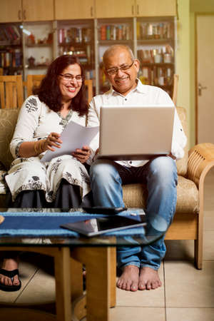 Senior Indian/asian couple accounting, doing home finance and checking bills with laptop, calculator and money while sitting on sofa/couch at homeの写真素材