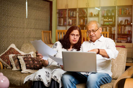 Senior Indian/asian couple accounting, doing home finance and checking bills with laptop, calculator and money while sitting on sofa/couch at homeの写真素材
