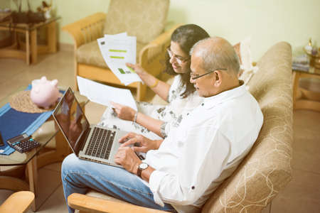 Senior Indian/asian couple accounting, doing home finance and checking bills with laptop, calculator and money while sitting on sofa/couch at homeの写真素材