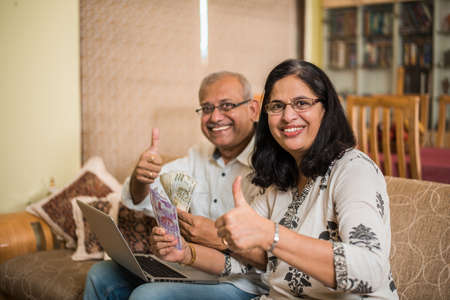 Senior Indian/asian couple accounting, doing home finance and checking bills with laptop, calculator and money while sitting on sofa/couch at homeの写真素材