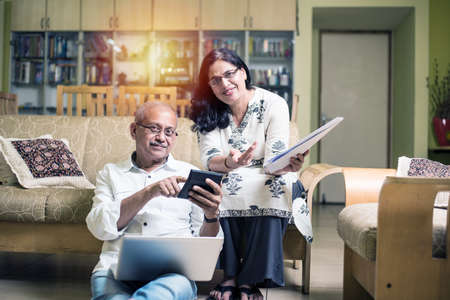 Senior Indian/asian couple accounting, doing home finance and checking bills with laptop, calculator and money while sitting on sofa/couch at homeの写真素材