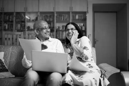 Senior Indian/asian couple accounting, doing home finance and checking bills with laptop, calculator and money while sitting on sofa/couch at homeの写真素材