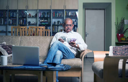 senior Indian/asian man accounting with money, laptop and calculator at homeの写真素材