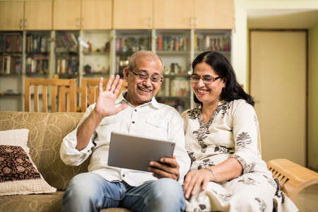 Indian/asian Senior couple video chatting on tablet/computer while sitting at couch or in garden at home, selective focusの写真素材