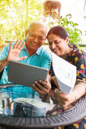 Indian/asian Senior couple video chatting on tablet/computer while sitting at couch or in garden at home, selective focusの写真素材