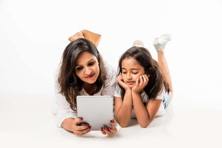Indian mother and daughter lying on floor with book, laptop or tablet computer studying or readying...