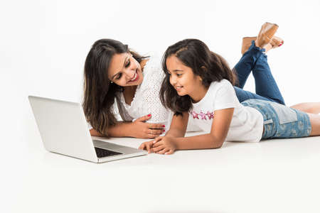 Indian mother and daughter lying on floor with book, laptop or tablet computer studying or readying...