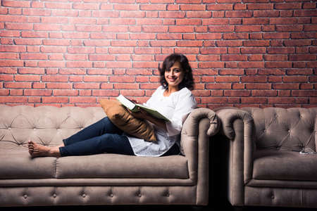 Indian lady reading book while sitting over couch/sofa, having coffee