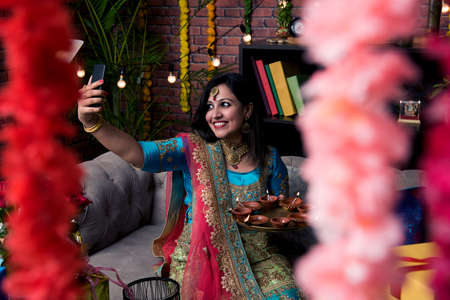 Indian girl holding diwali diya in traditional wear, clicking selfie picture or speaking over smartphone.の写真素材