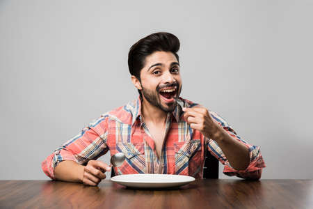 empty plate and Indian man with beard holding spoon and fork, wearing checkered shirt and sitting at tableの写真素材