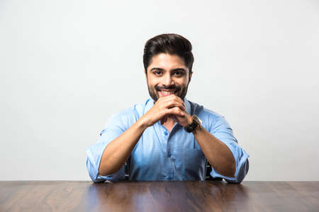 Portrait of Indian Male businessman sitting at table / desk in office, over grey background