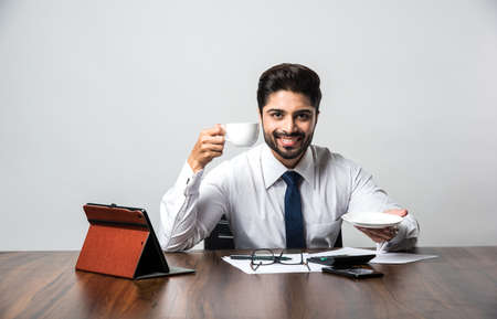 Indian businessman having coffee / tea at work / office, sitting at desk or table holding cup and saucerの写真素材