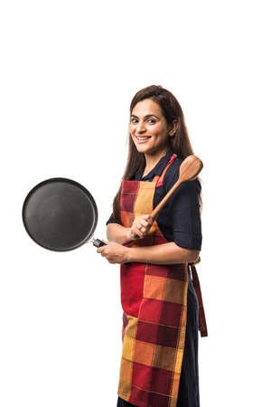 Indian / asian woman chef wearing Apron and holding Pan and spatula while standing isolated over white backgroundの写真素材
