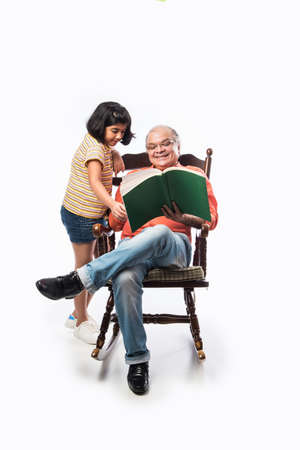 Indian Senior man or grandpa reading book with granddaughter or grand daughter while sitting on rocking chair against white backgroundの写真素材