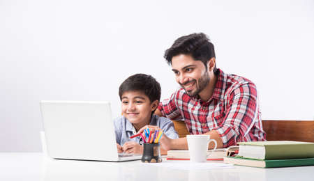 Cute Indian boy with father or male tutor doing homework at home using laptop and books - online...