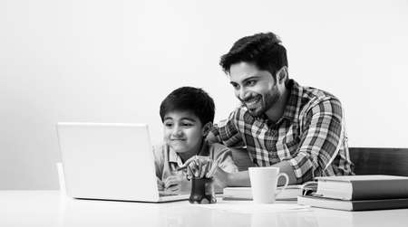 Cute Indian boy with father or male tutor doing homework at home using laptop and books - online...