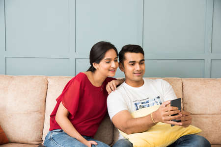Indian asian young couple using smartphone together while sitting on sofa or couch and modern interior space or homeの写真素材