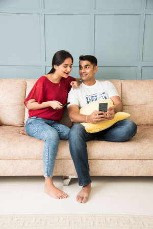Indian asian young couple using smartphone together while sitting on sofa or couch and modern interior space or homeの写真素材
