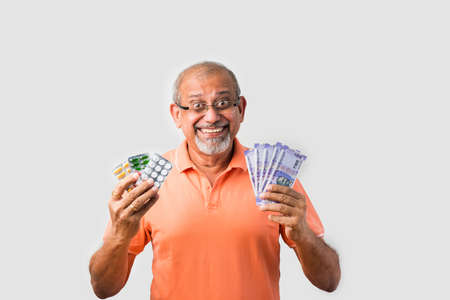 Senior retired Old Indian asian man showing medicine blistep pack of tablets with currency or mobile...