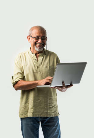 Indian asian senior adult man or grandpa using laptop and holding paper currency notes against white backgroundの写真素材