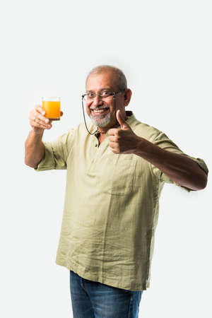 Indian asian senior or old man drinking fresh orange juice in a glass while standing against white backgroundの写真素材