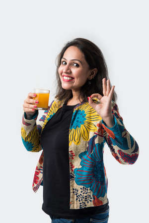 Indian asian young woman or girl drinking fresh orange juice in a glass while standing against white...