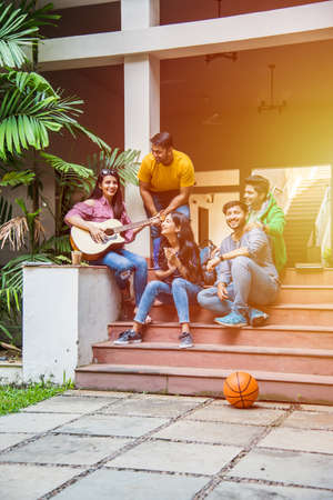 Asian Indian college students playing music with guitar while sitting in campus on stairs or over...