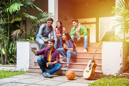 Asian Indian college students taking selfie picture using smartphone in campus while sitting on...