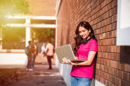 Asian Indian college student in focus working on laptop or reading book while other classmates in the background, outdoor picture in university campusの写真素材