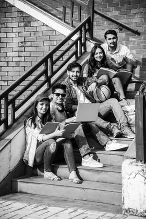 Young Asian Indian college students reading books, studying on laptop, preparing for exam or working on group project while sitting on grass, staircase or steps of college campusの写真素材