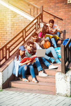 Young Asian Indian college students reading books, studying on laptop, preparing for exam or working on group project while sitting on grass, staircase or steps of college campusの写真素材