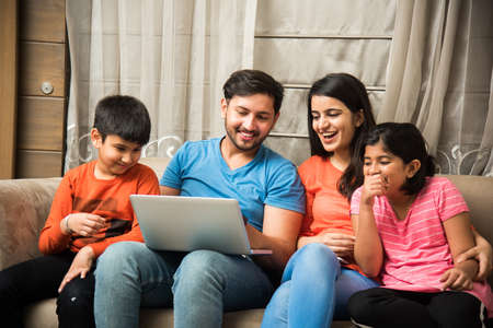 Indian family sitting on sofa and using smartphone, laptop or tablet, watching movie or surfing...