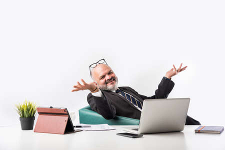 Portrait of a satisfied senior Indian Asian businessman sitting at workplace in front of the computer and looking at the camera while relaxing. Old professional man feet on office deskの写真素材