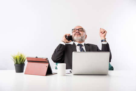 Asian Indian senior financial businessman sitting at his workstation or desk in front of a computer,...