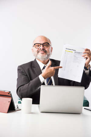 Asian Indian senior financial businessman sitting at his workstation or desk in front of a computer, laptop and tablet. Speaking on phone while doing some paperworkの写真素材