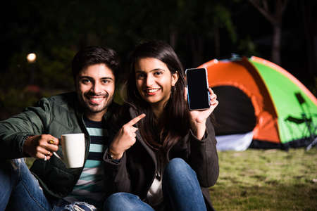 Happy young asian indian couple camping, sitting by tent at campsite relaxing and drinking tea.の写真素材