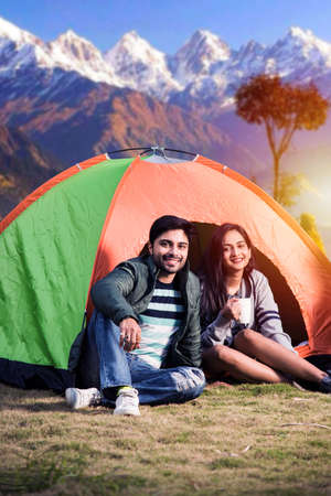 Happy young asian indian couple camping, sitting by tent at campsite relaxing and drinking tea.