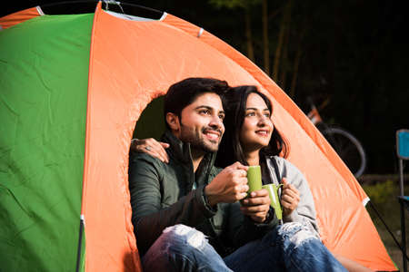 Happy young asian indian couple camping, sitting by tent at campsite relaxing and drinking tea.の写真素材