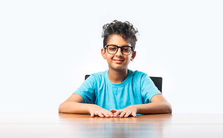 Indian asian boy or male child sitting at table or desk, looking at cemara, thinking or pointing somethingの写真素材
