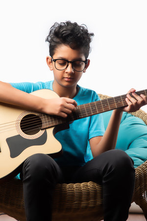 Indian asian boy playing acaustic guitar while sitting against white background or brick wala on chairの写真素材