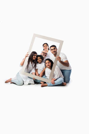 Happy Indian asian multigenerational family of six looking through an empty frame, standing or sitting against white background wearing white cloths and blue denim jeansの写真素材
