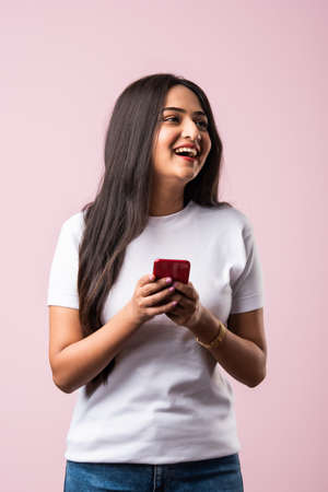 Pretty millennial indian girl isolated on pink studio background text messaging on cell phone or using smartphone with wireless connectionの写真素材