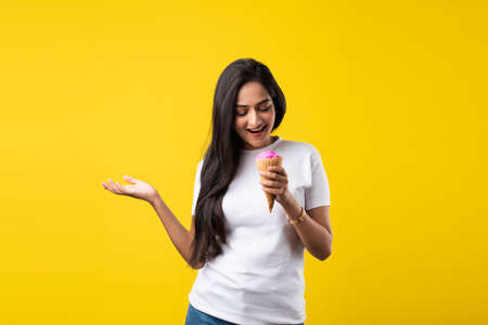 Pretty Indian girl or young Asian woman eating strawberry ice cream in cone against yellow studio...
