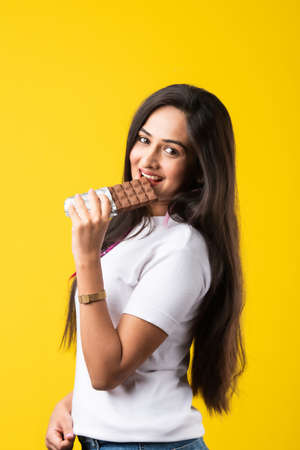 Portrait of a happy young Indian asian young woman biting chocolate bar isolated over pink or yellow...