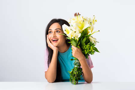 Close up portrait of an attractive young Indian woman holding flower bouquet while sitting isolated at table or desk against white backgroundの写真素材