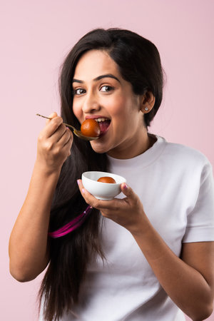 Pretty Indian young girl or woman eating sweet Gulab Jamun against pink background